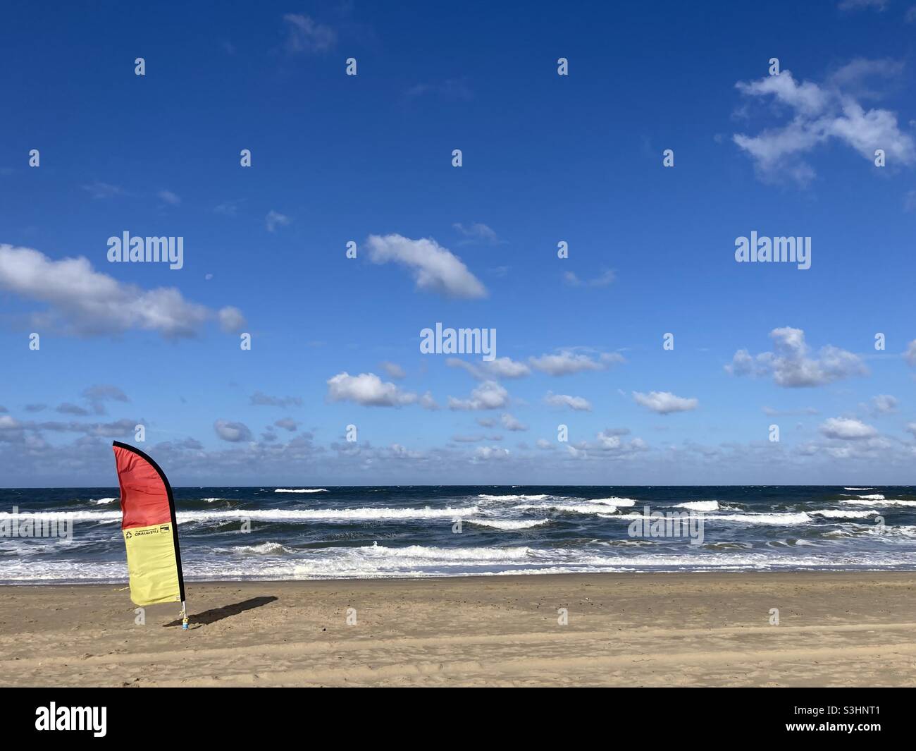 A red yellow flag stands on the beach by the sea in a strong wind in sunny weather and blue skies - Smartphone Captured Stock Image