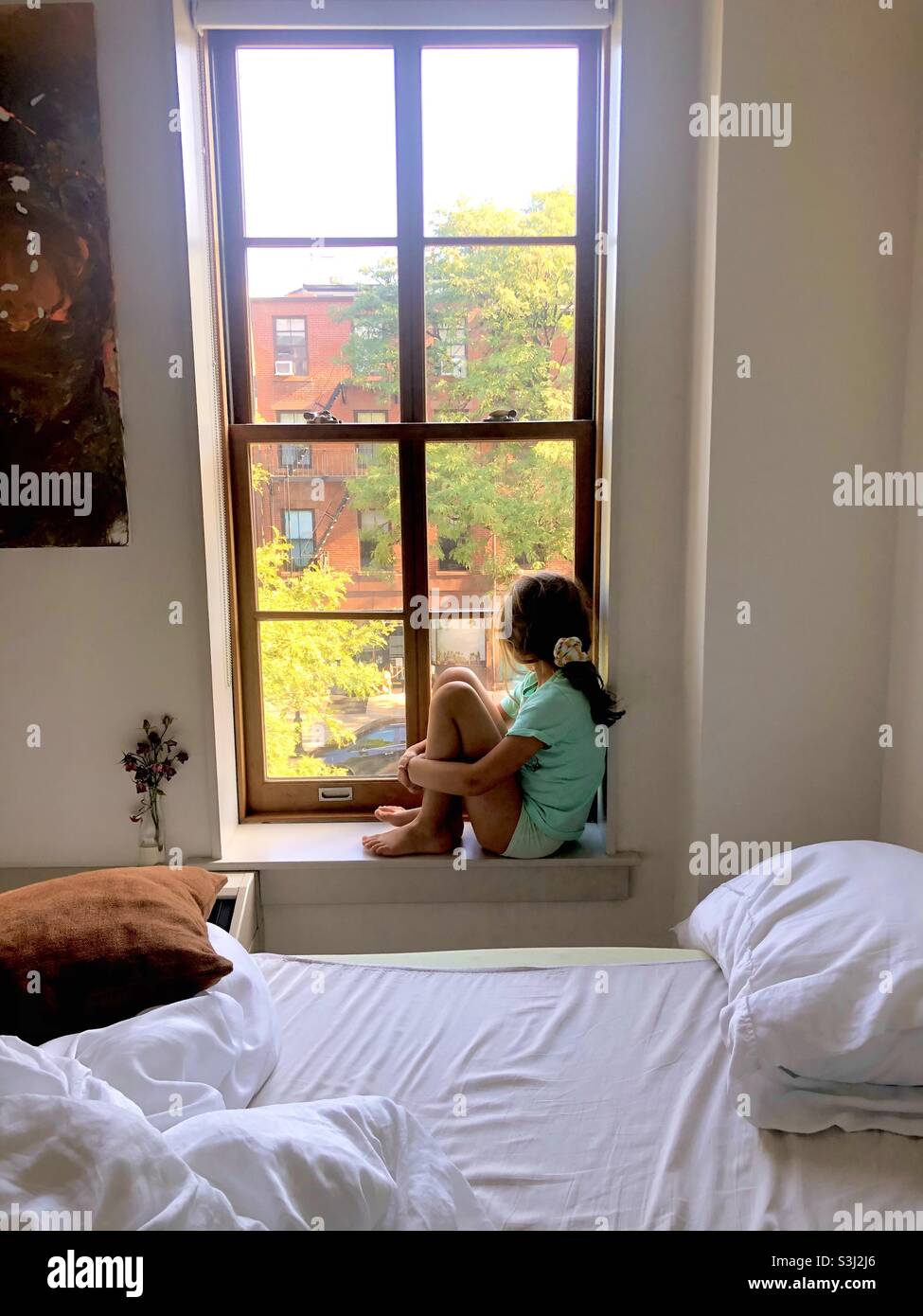 Eight year old girl sitting on the window sill in an urban setting and looking out the window. - Smartphone Captured Stock Image