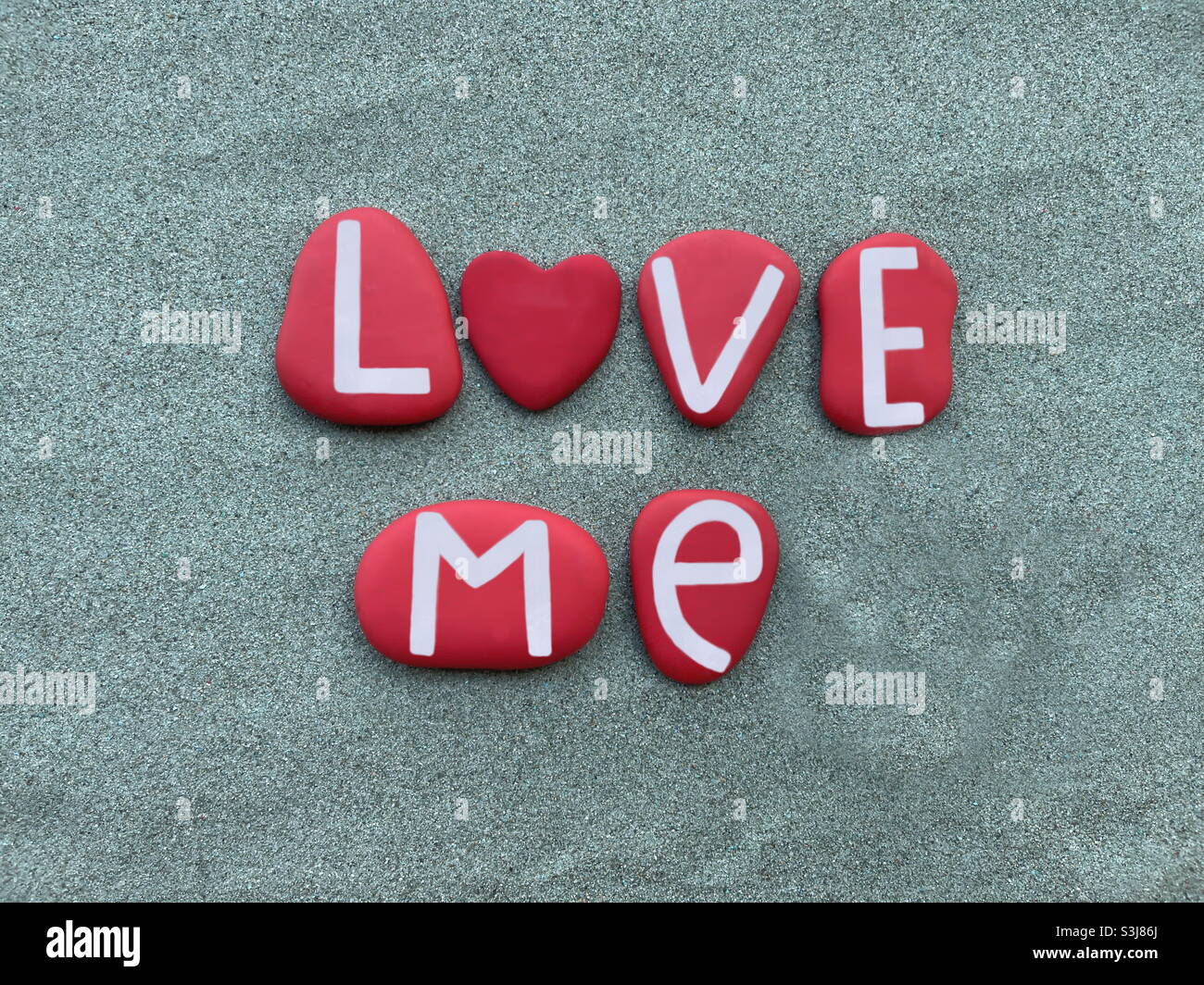 Love me text composed with red colored stone letters and a red painted stone heart over green sand Stock Photo