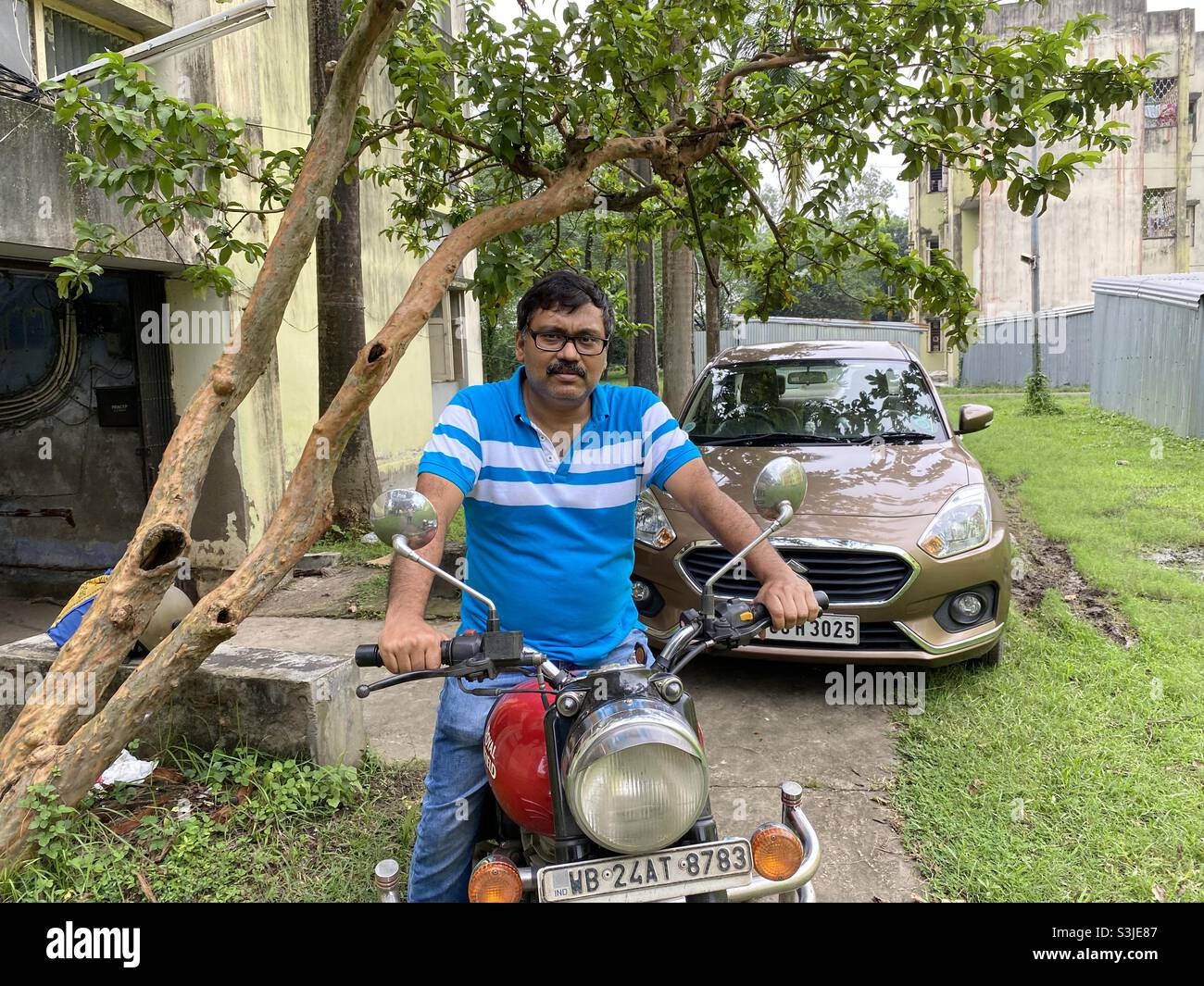 Middle aged Indian man sit on a two wheelers in a residential area of Bengal with four wheelers at background - Smartphone Captured Stock Image