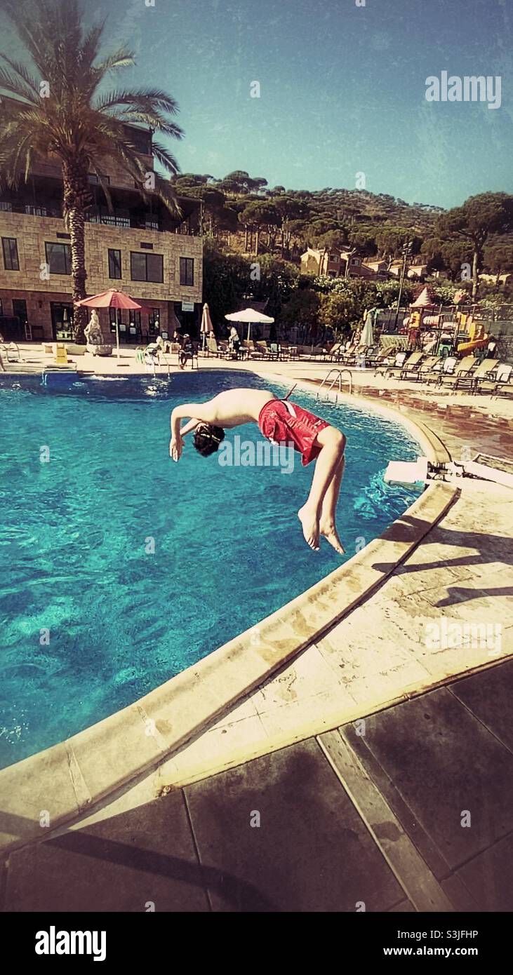 13 years old boy jumping backflip in outdoors swimming pool wearing red swimming shorts Stock Photo