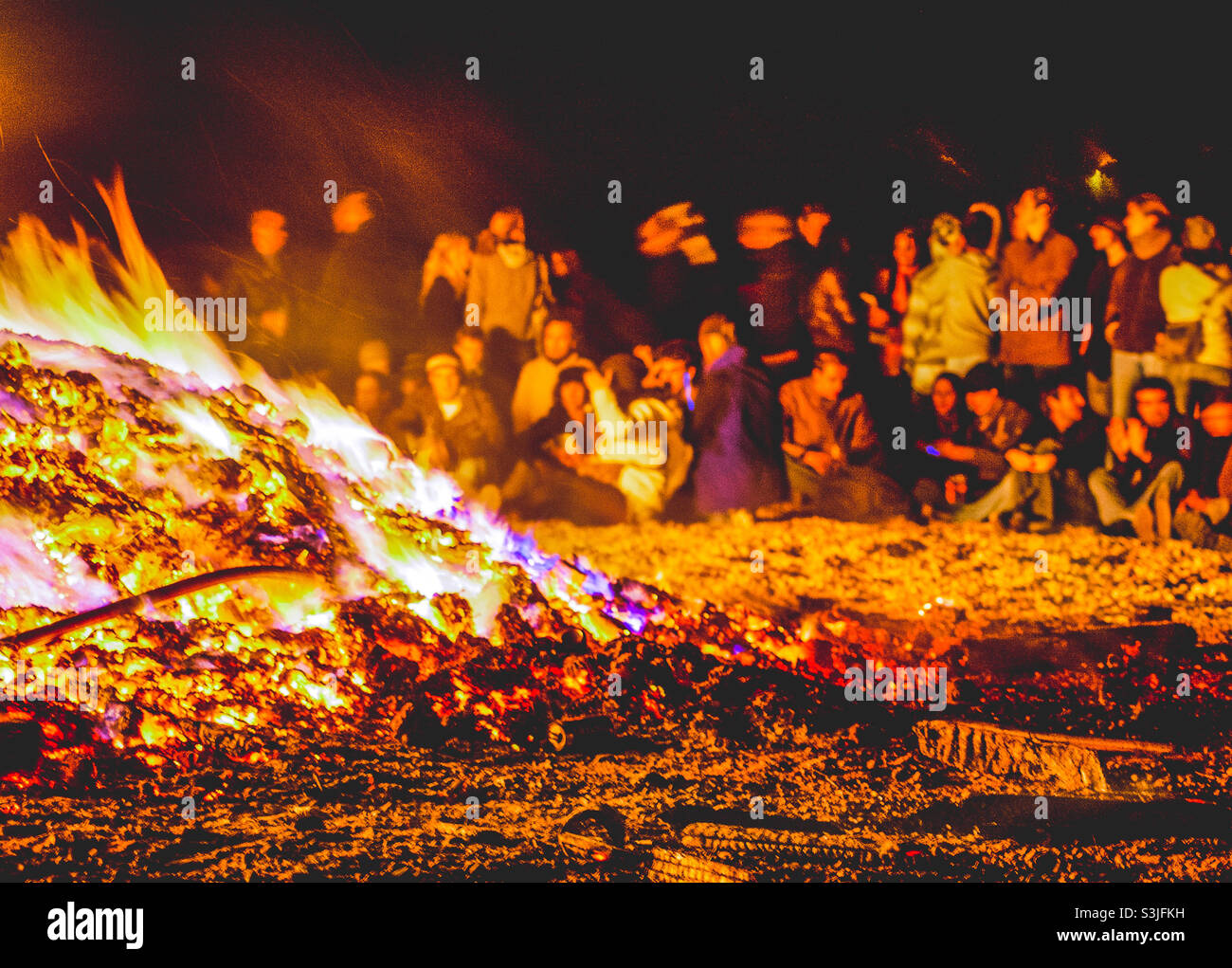 A blurred crowd of people can be seen on the beach in the background next to a large and very hot bonfire - Smartphone Captured Stock Image