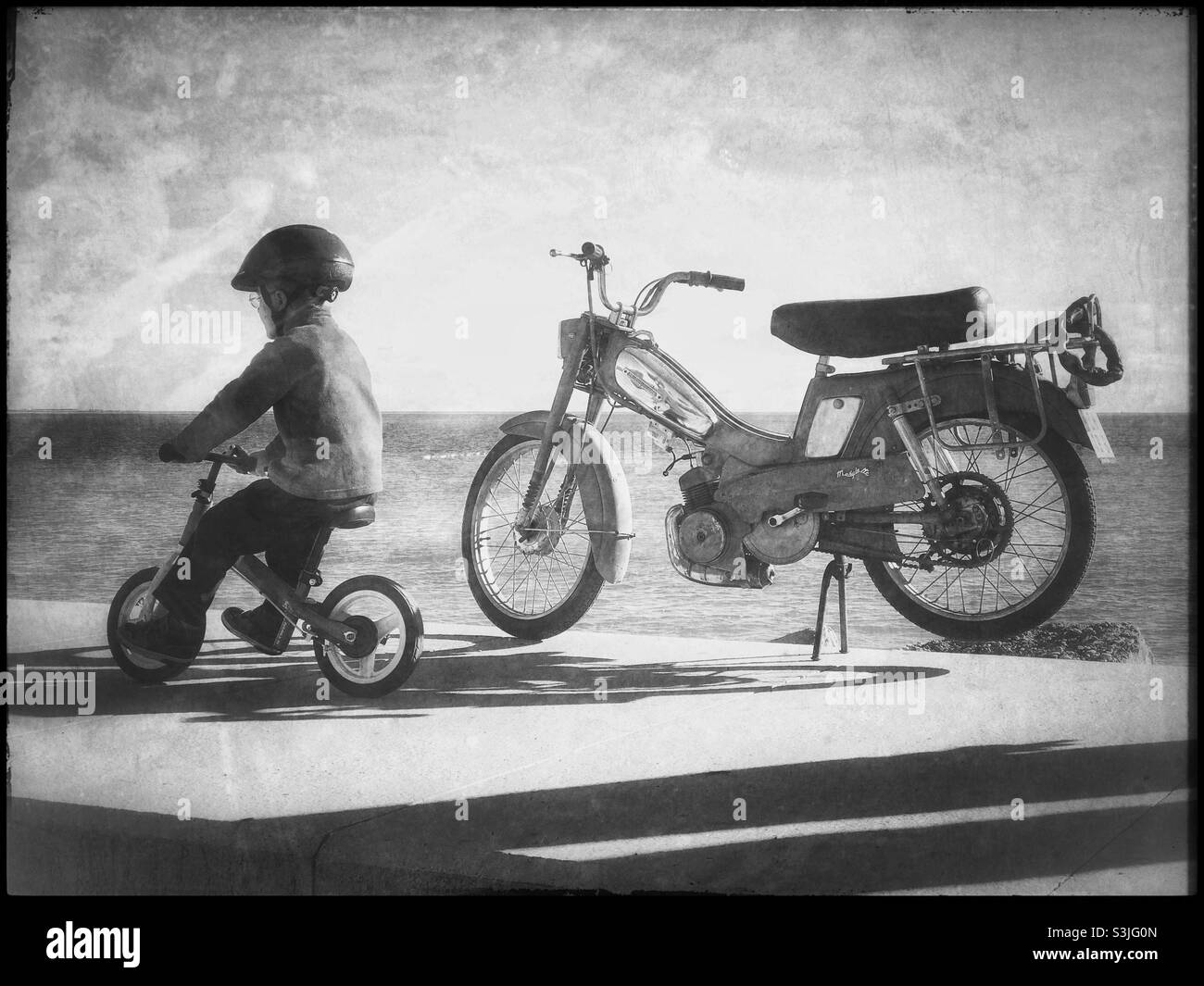 A boy rides past a motorcycle on his balance bike. A humorous visual play on words? Dreaming of adulthood. Where’s Dad? Copy space. Free to enjoy life riding around? What’s does the future hold? ©️ CH - Smartphone Captured Stock Image