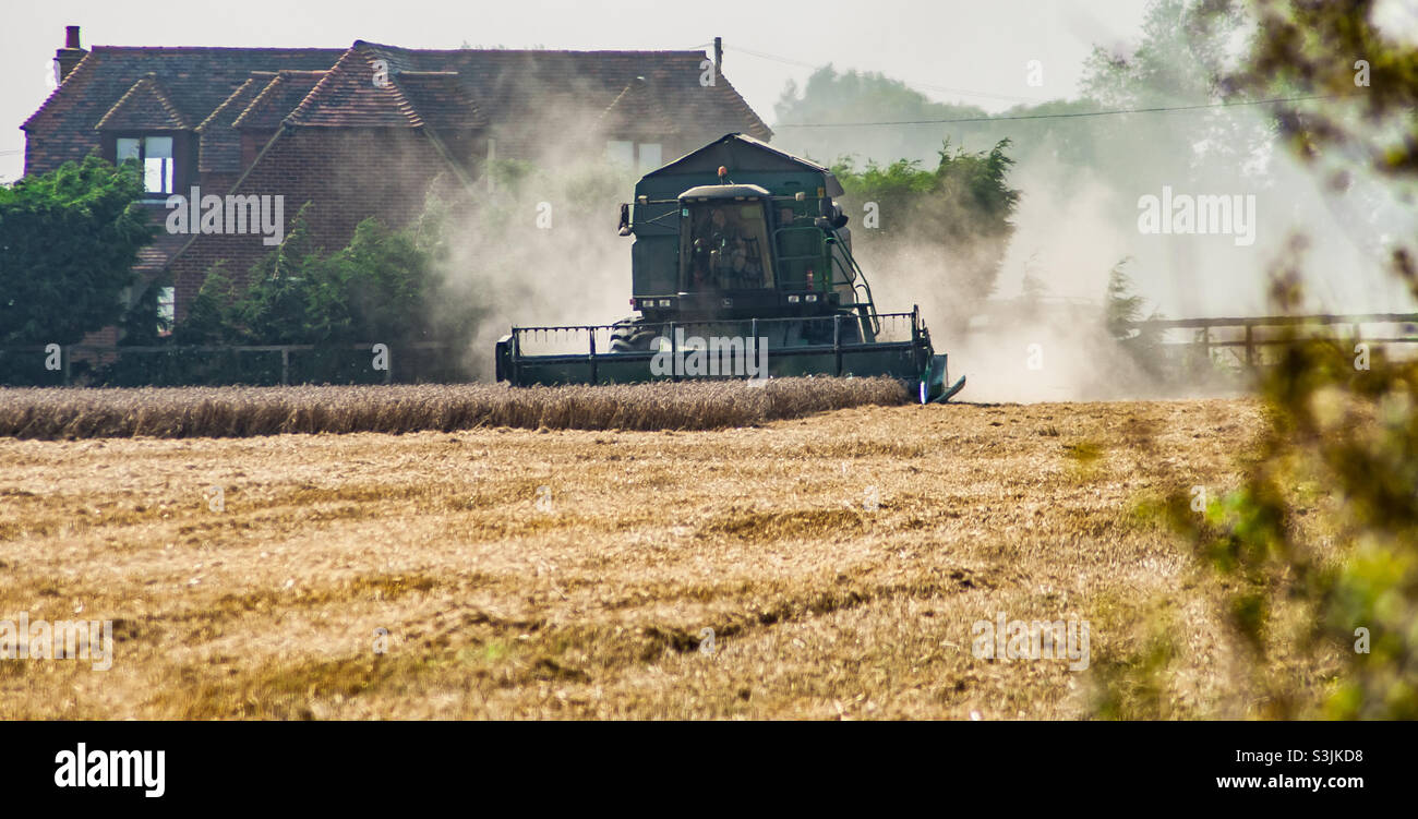 A harvester works the field, creating much dust in the air, in front of an old farmhouse - Smartphone Captured Stock Image