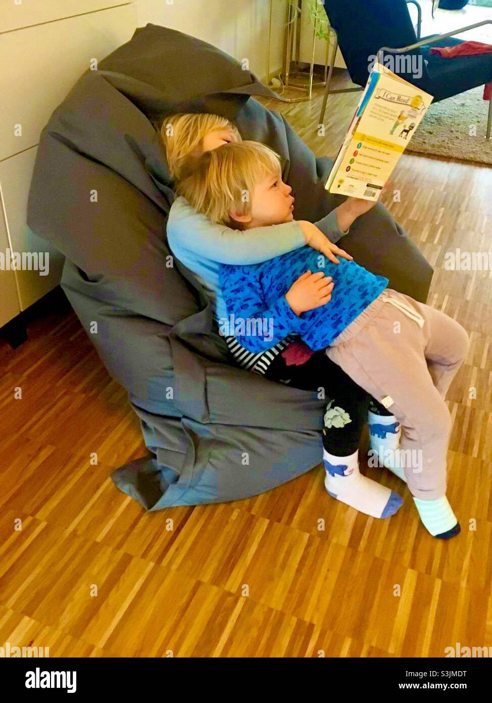 To blonde children sitting on top of each other in a bean bag chair reading - Smartphone Captured Stock Image