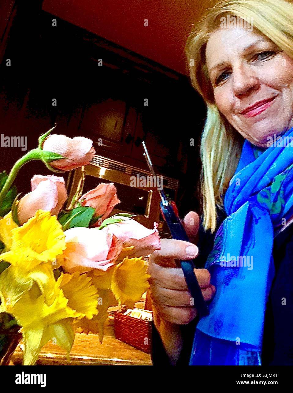 A blonde woman in a bright blue scarf holding scissors to cut a bouquet of pink roses and yellow daffodils - Smartphone Captured Stock Image
