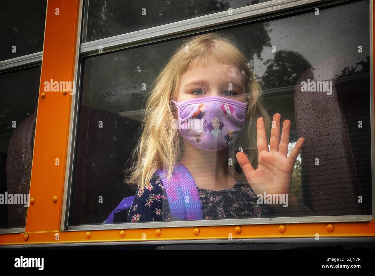 Masked blonde haired school girl waving from bus window on the first day of school Stock Photo