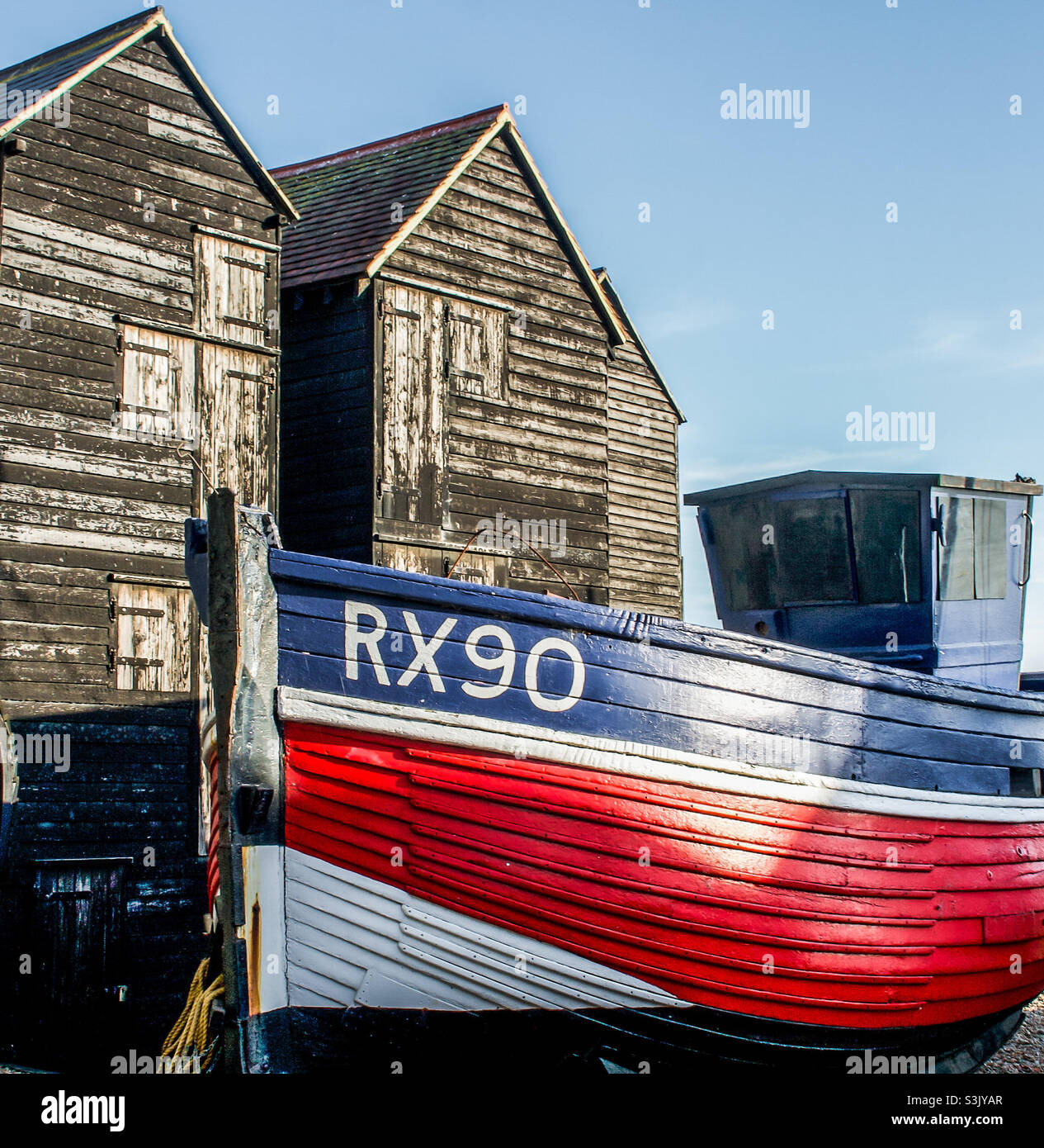 A blue, red and white fishing vessel sits in front of the tall, black, wooden huts in Hastings UK - Smartphone Captured Stock Image