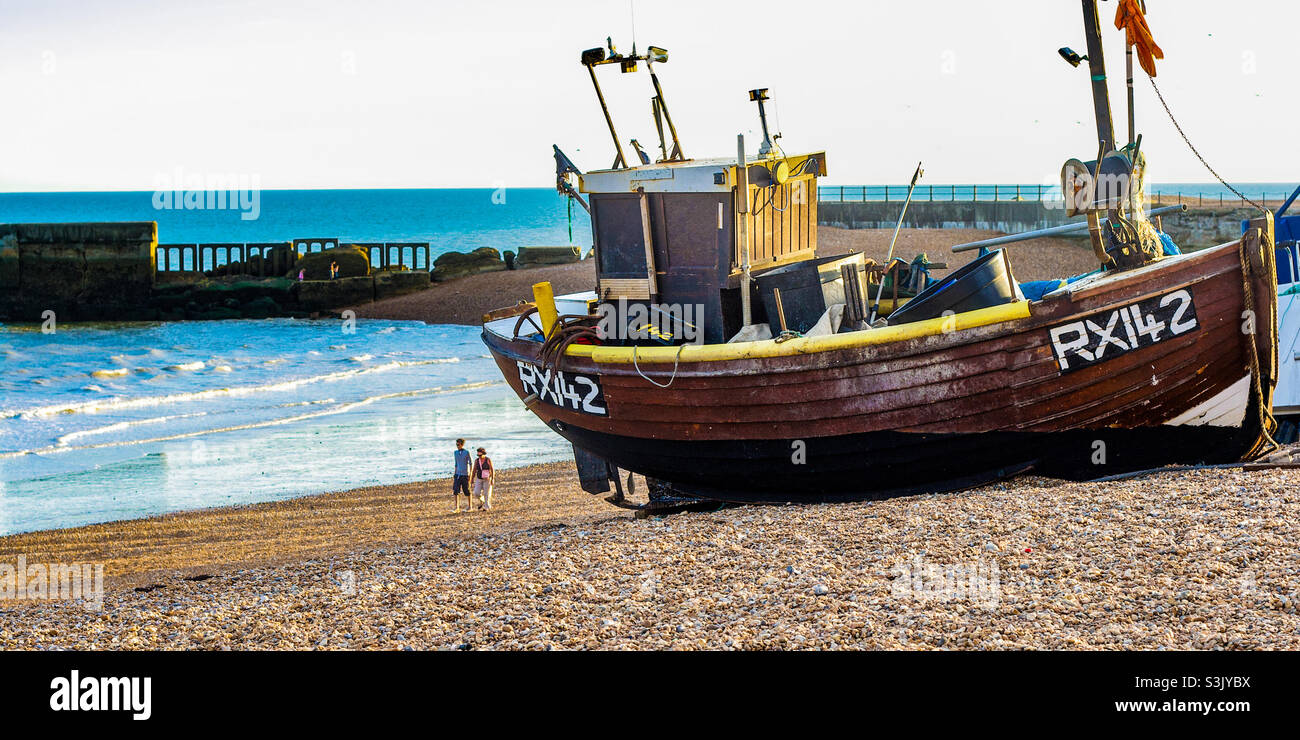 A fishing vessel rests on a stony beach, 2 people can be seen in the distance walking near the old harbour arm - Smartphone Captured Stock Image