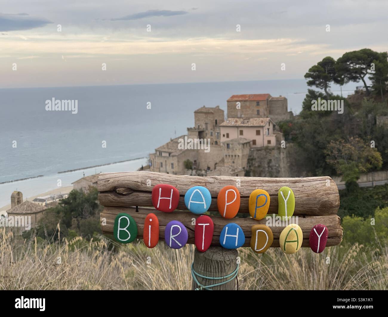 Creative Happy Birthday with colored stone letters front of a medieval village with sea background - Smartphone Captured Stock Image