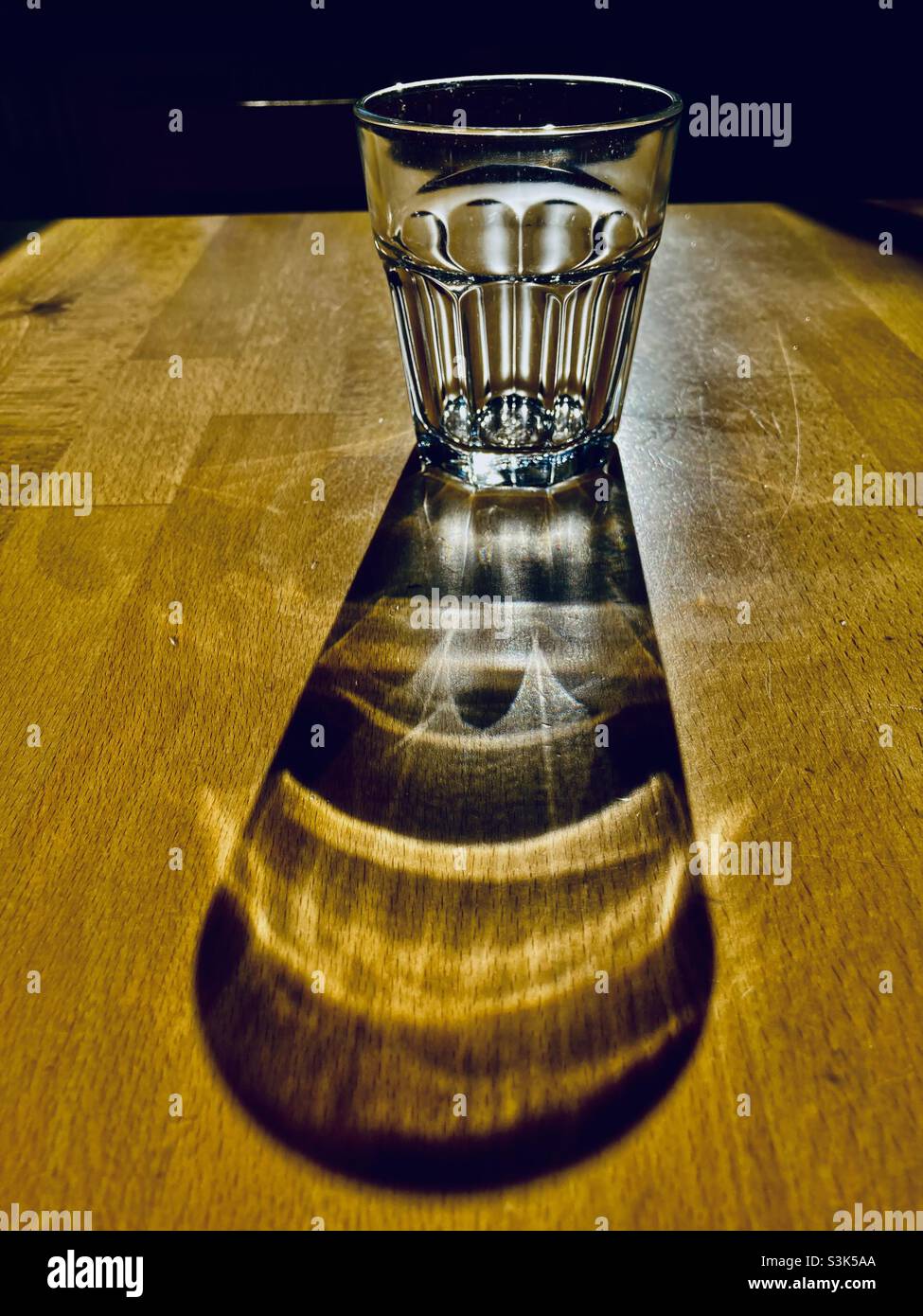 a glass of water stands on a wooden table and casts a long shadow - Smartphone Captured Stock Image