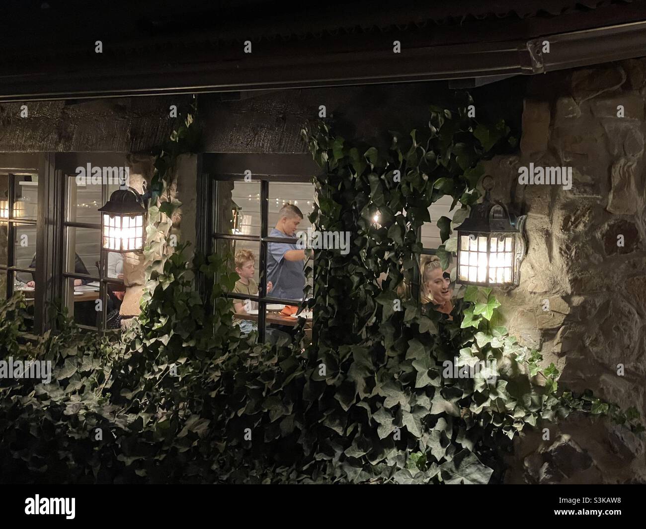An ivy covered storefront is illuminated at dusk at Gardner Village in Utah, USA during its’ annual “WitchFest” each October. You can see shoppers and patrons milling about through the shop window. - Smartphone Captured Stock Image