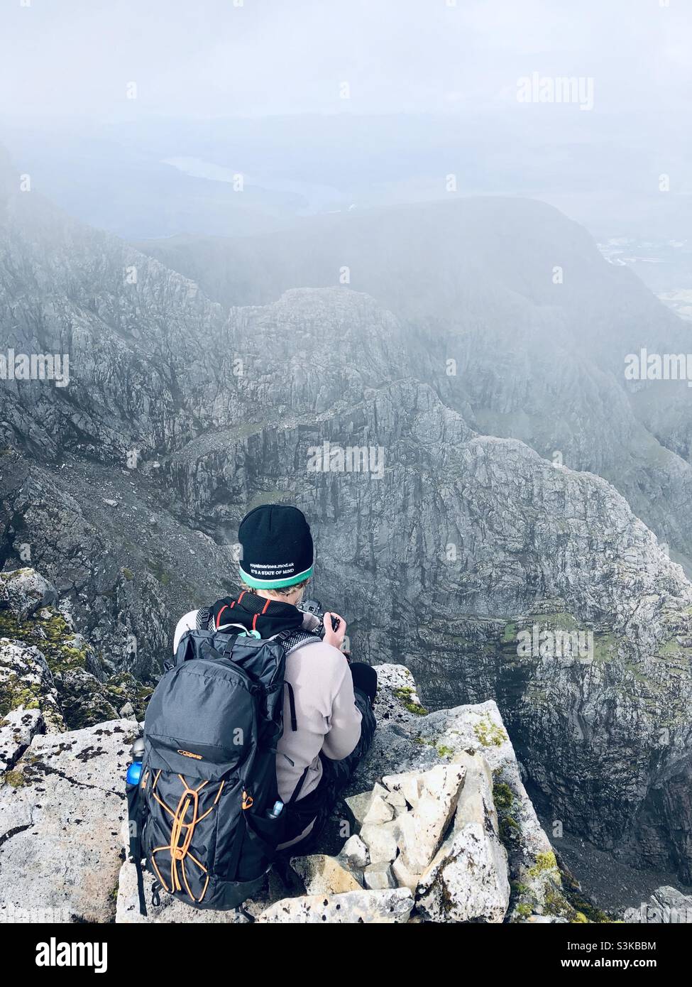 A person with a backpack sits on the edge of a cliff looking at the view of the mountains - Smartphone Captured Stock Image