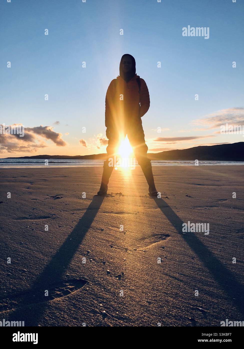 A person stands on the beach with the sun setting between their open legs in Scotland - Smartphone Captured Stock Image