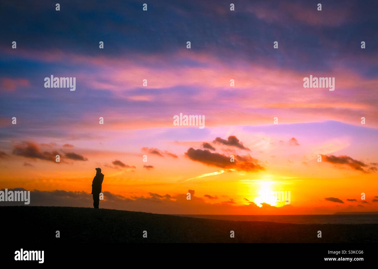 A vivid winter sunset silhouettes a lone man in warm clothing as he pauses during his evening stroll on the beach Stock Photo
