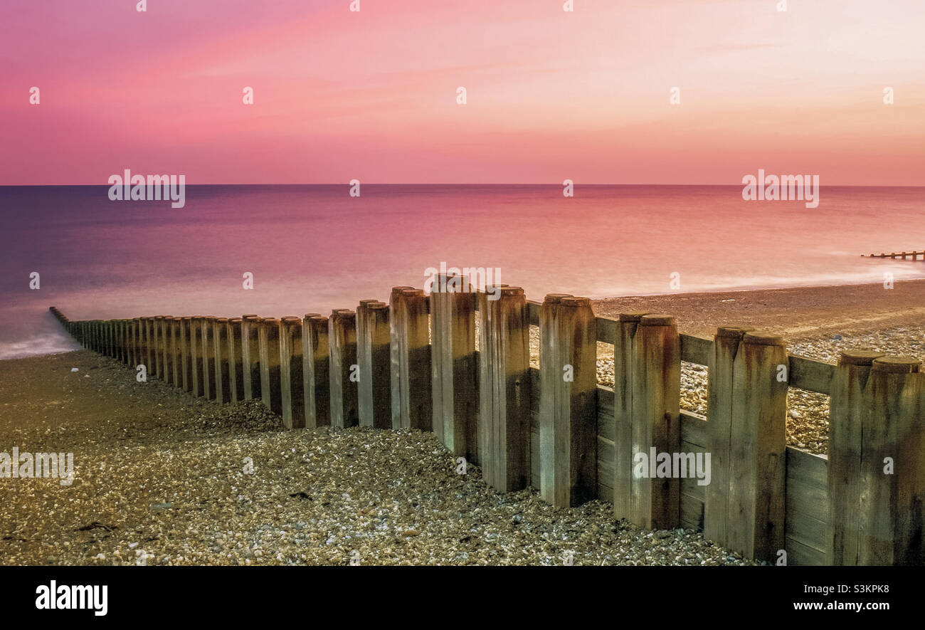 A vibrant pink and purple sky dominates the horizon on the sea, in the foreground wooden groynes lead down the beach to the shore - Smartphone Captured Stock Image