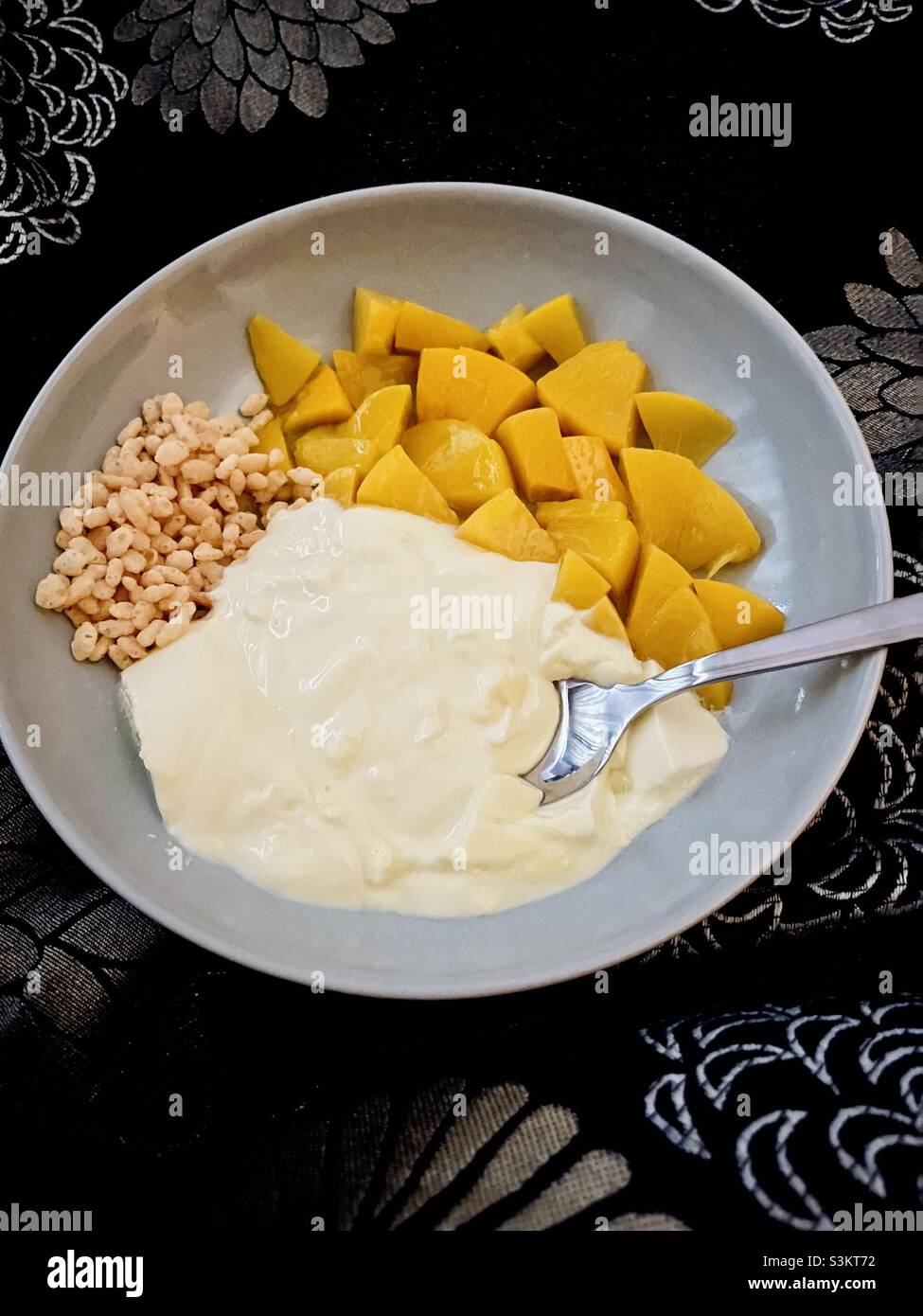 Breakfast in bed - yoghurt, chopped fruit and cereal in a bowl with a spoon on a black pattered duvet cover Stock Photo
