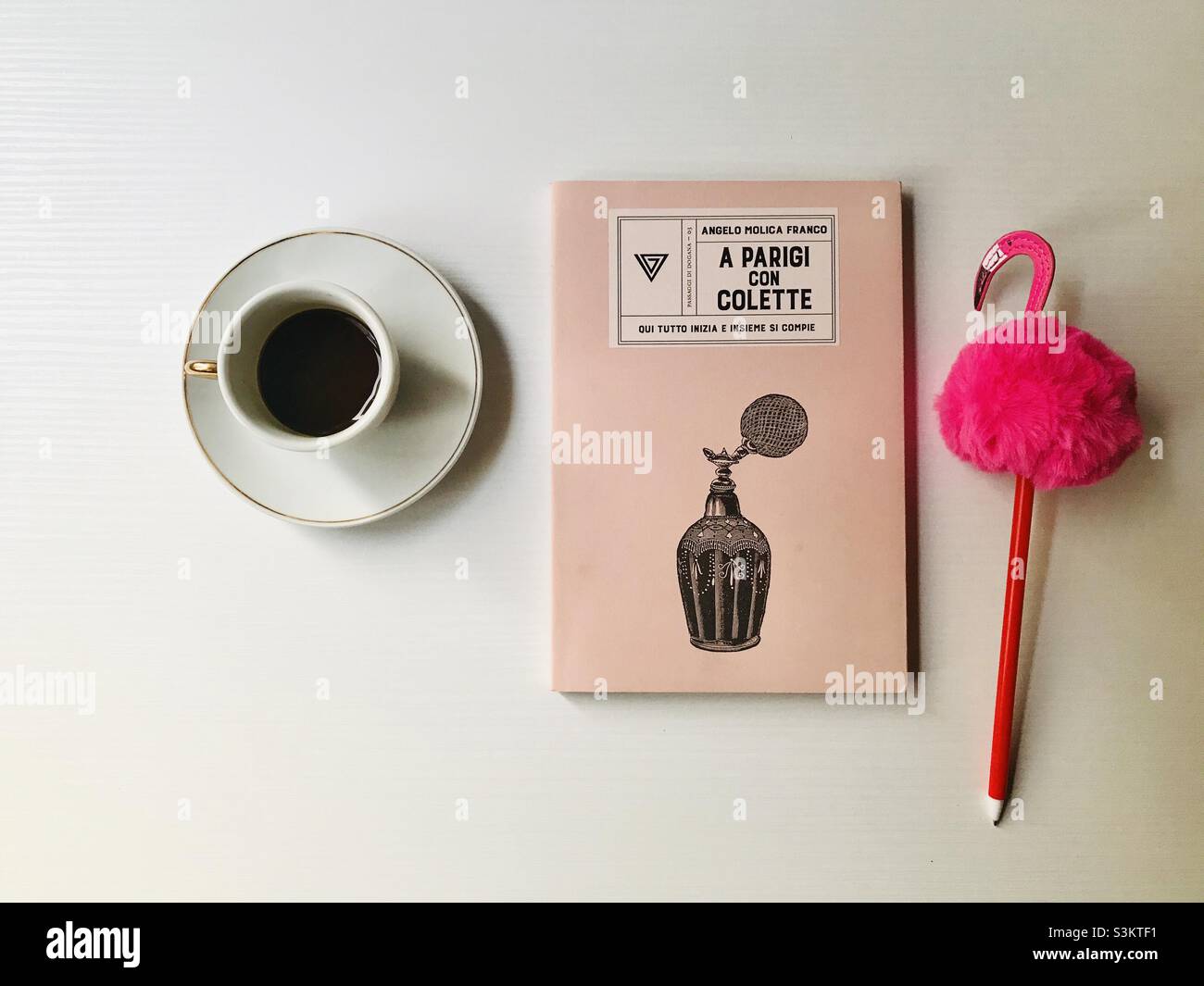 Close up from above of a cup of espresso coffee, a pink book about Paris and Colette and a pink flamingo pen on a white background. Stock Photo