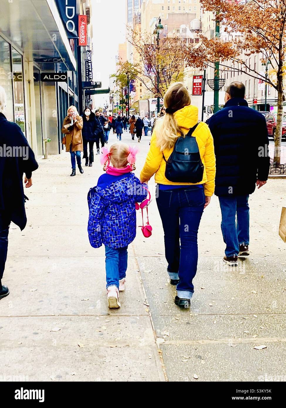 A cute little girl with pink ribbons in her hair walks with other pedestrians on a sidewalk in New York City Stock Photo