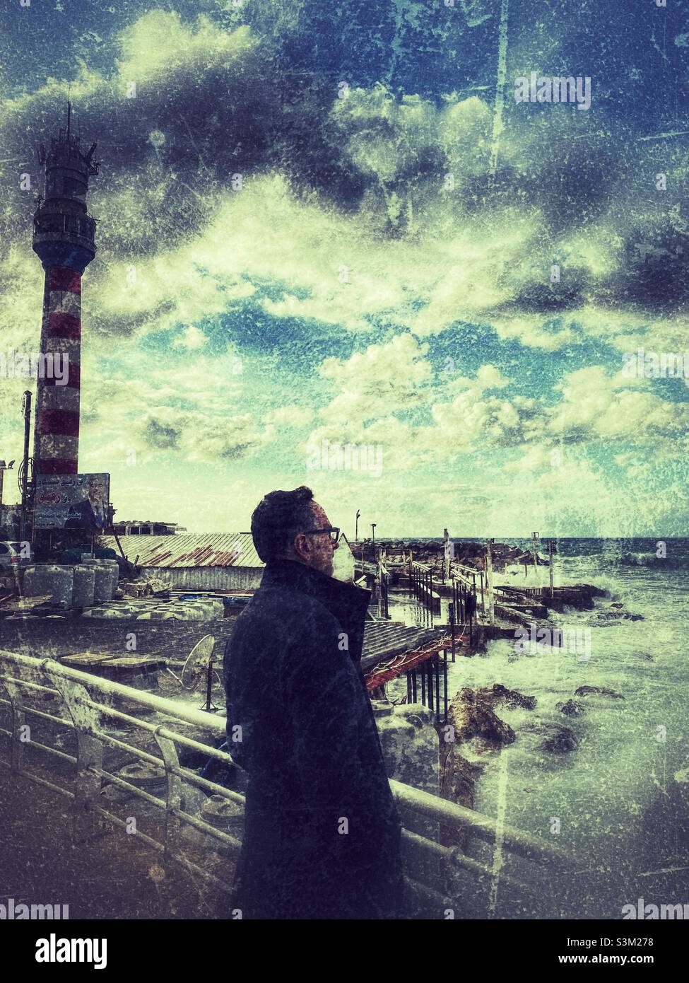 Middle eastern man with coat standing on the promenade watching sea waves - Smartphone Captured Stock Image