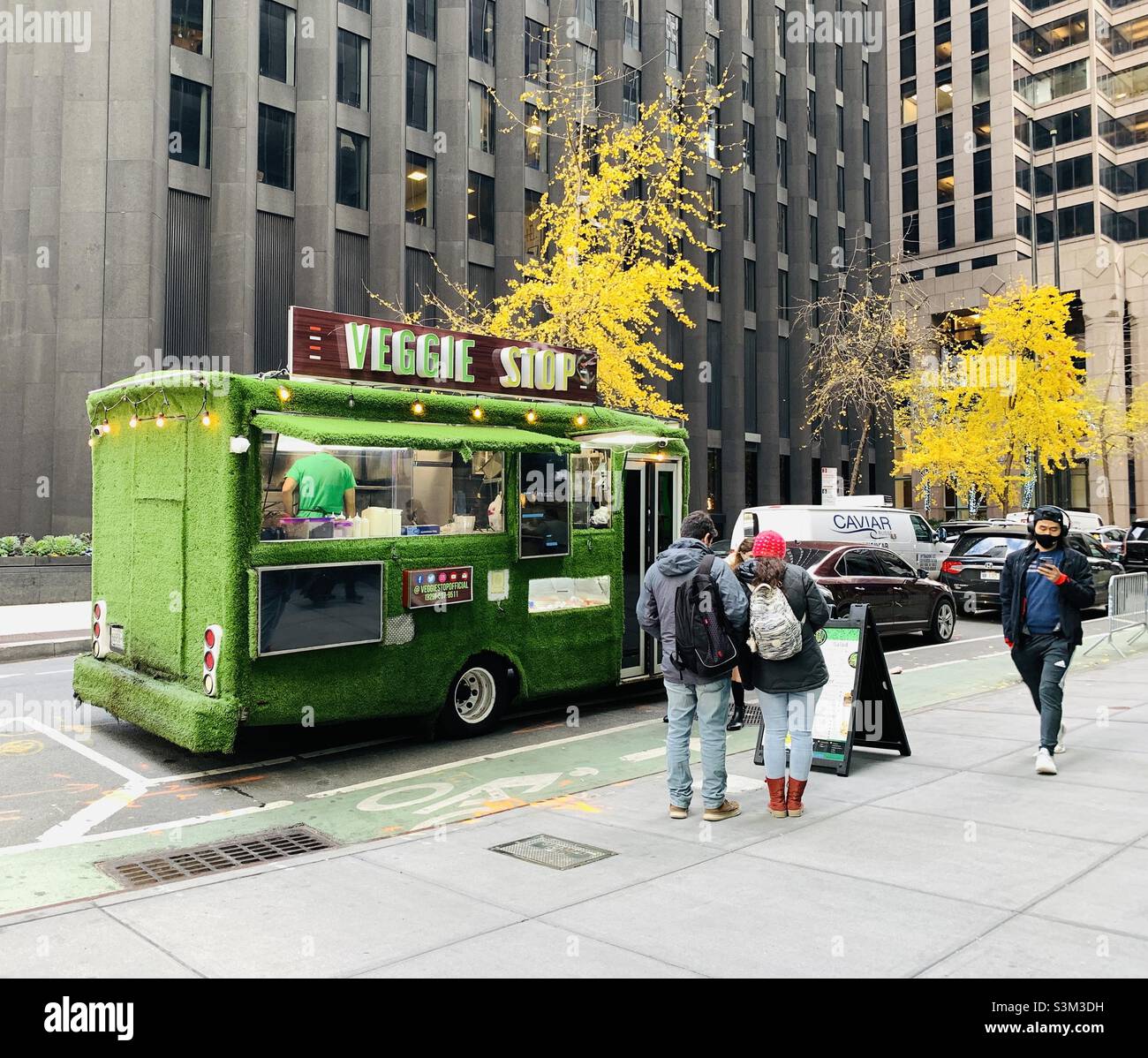 Three people looking the menu of veggie stop. One person walking by looking at his phone. - Smartphone Captured Stock Image