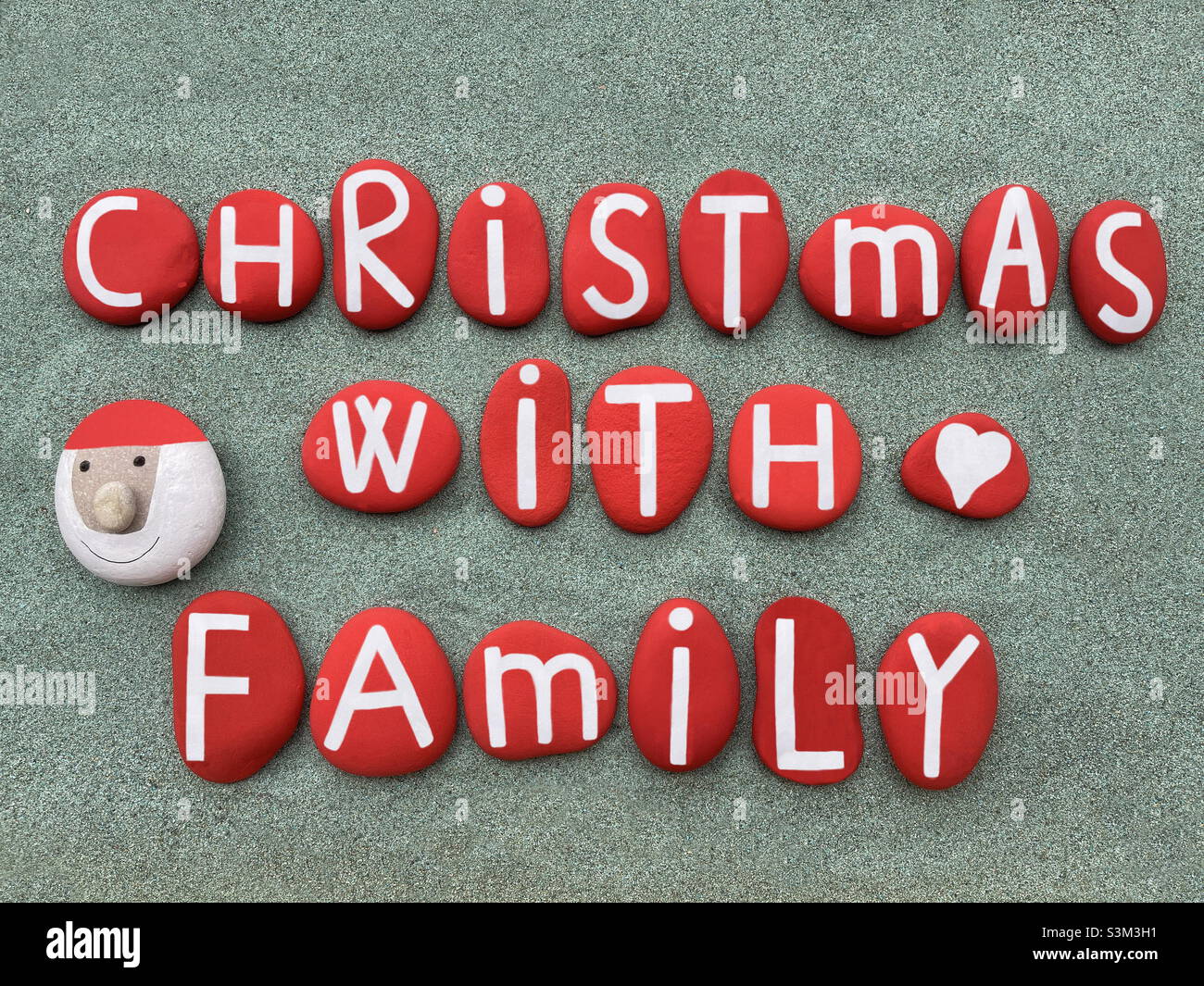 Christmas with family, creative text composed with red colored stone letters and a stone Santa Claus over green sand Stock Photo