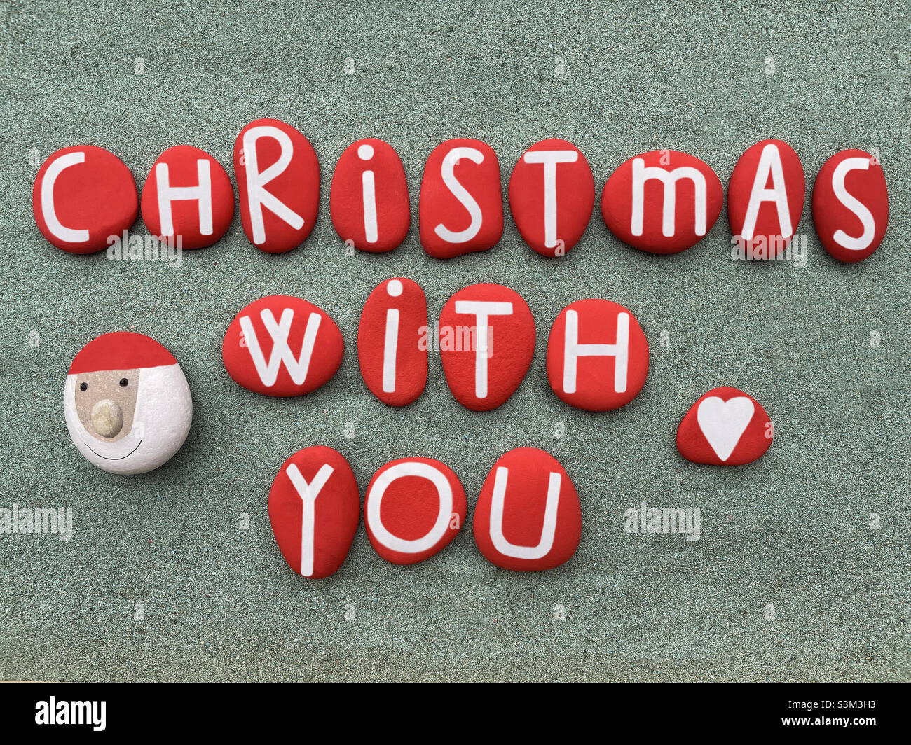 Christmas with you, creative phrase composed with hand painted red stone letters and s stone Santa Claus over green sand Stock Photo