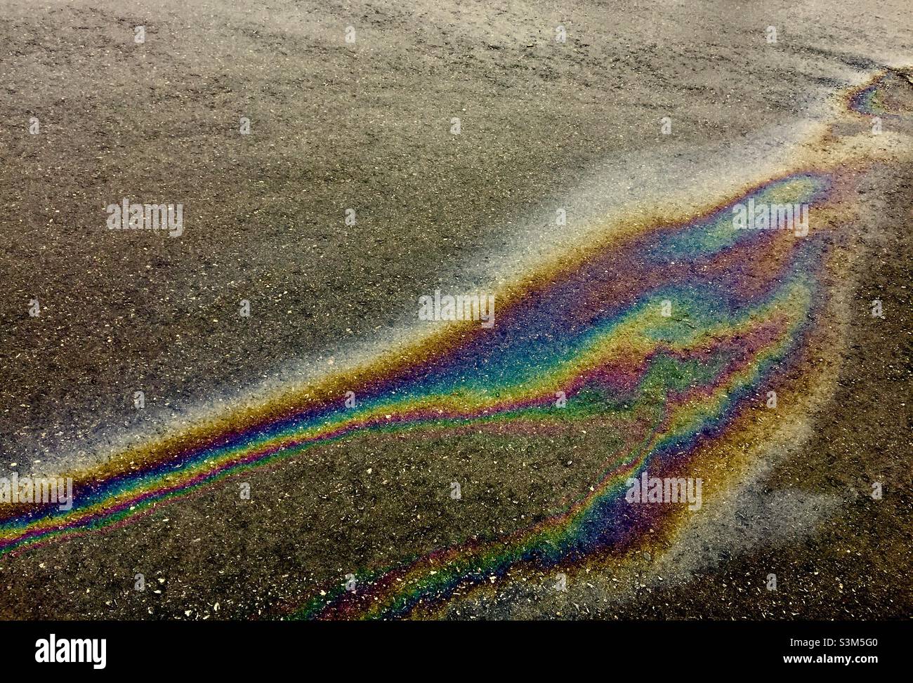 Rainbow colours on the road. Oil gasoline drip from a vehicle becomes a slow moving stream as rain falls on asphalt, Ontario, Canada - Smartphone Captured Stock Image