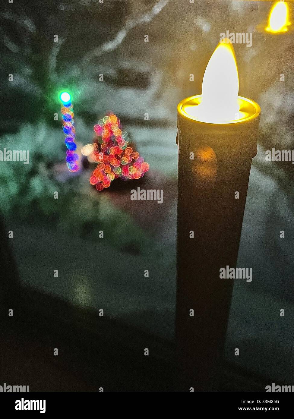 A battery operated candle glows and flickers in a home’s upstairs window, looking out into their front yard with shining Christmas lights and new snowfall in Utah, USA. - Smartphone Captured Stock Image
