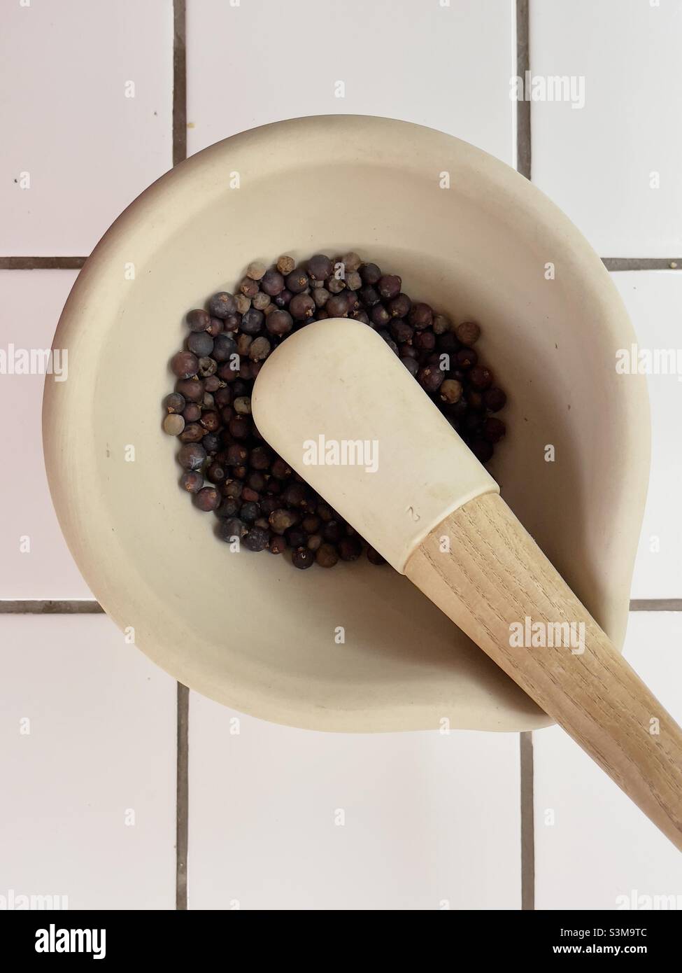 A pestle and mortar containing juniper berries resting on a white tiled worktop - Smartphone Captured Stock Image