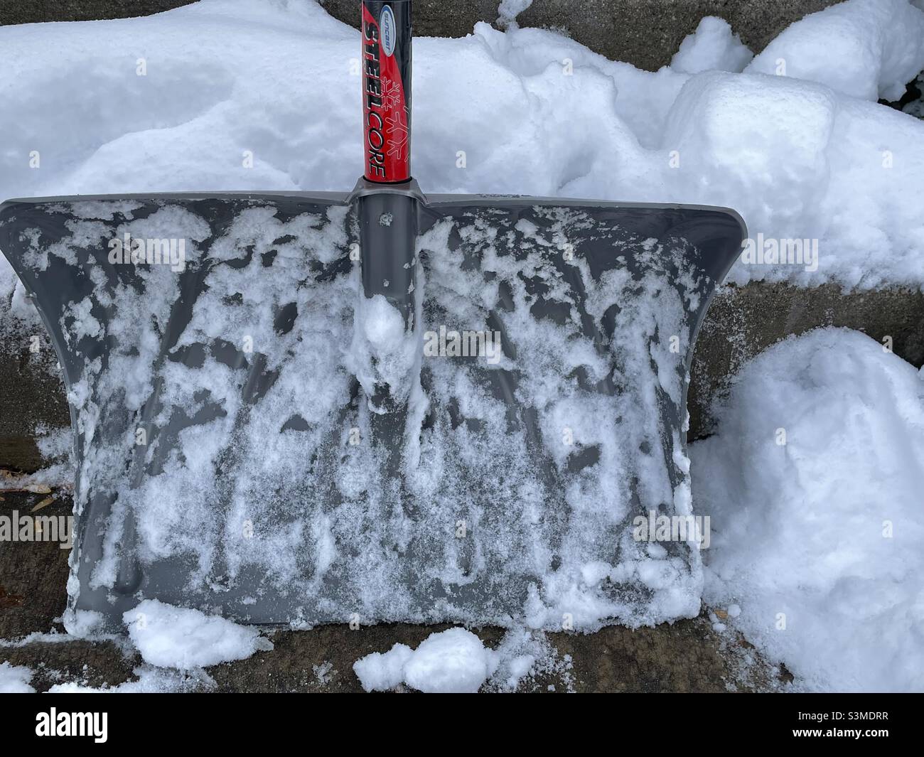 A foot of new snowfall necessitated the use of this heavy duty snow shovel in Utah, USA. The snowy shovel is propped against snow-covered porch steps to make this seasonal still life. - Smartphone Captured Stock Image