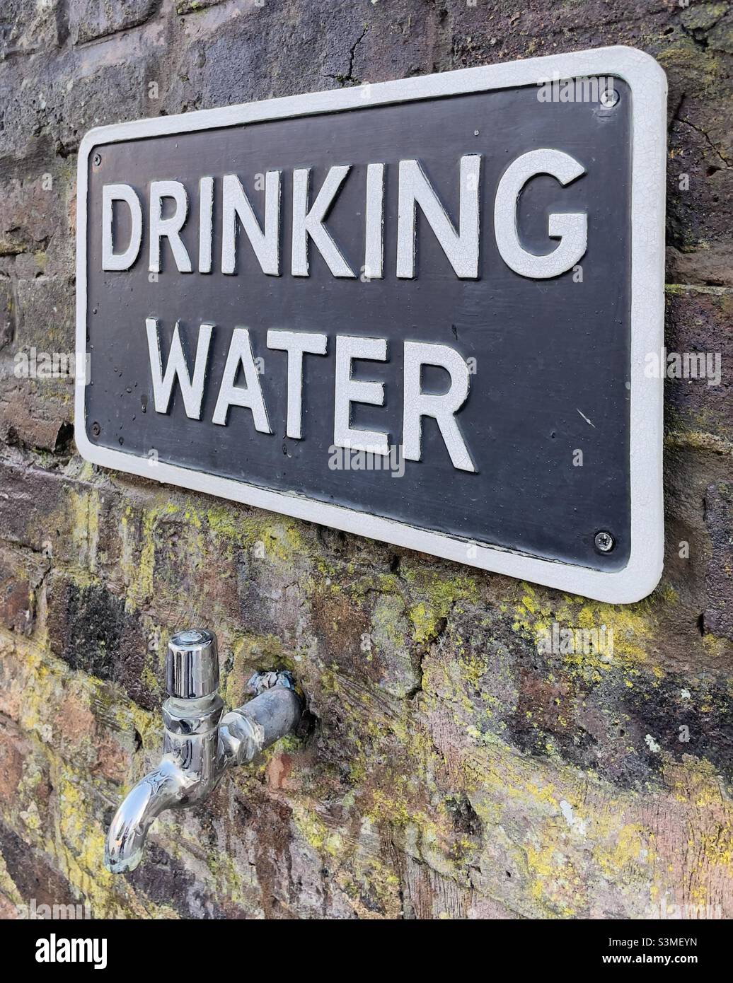 A tap projecting from a brick wall with a sign above it saying “drinking water” - Smartphone Captured Stock Image