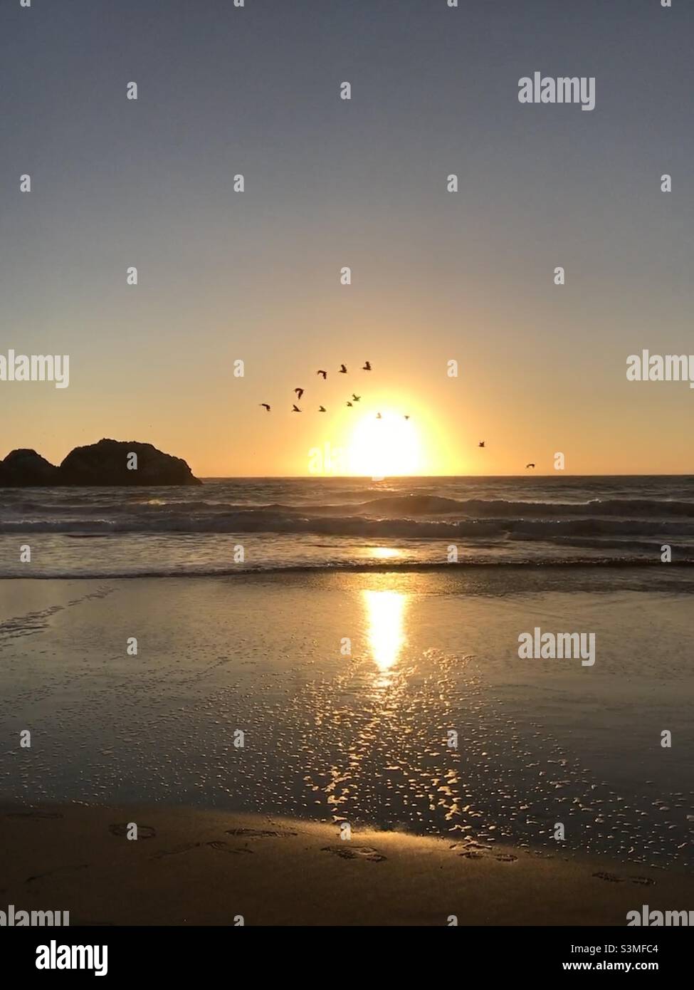 A bright Orange Sun setting over the ocean with birds flying past and a rock island in the background. Stock Photo