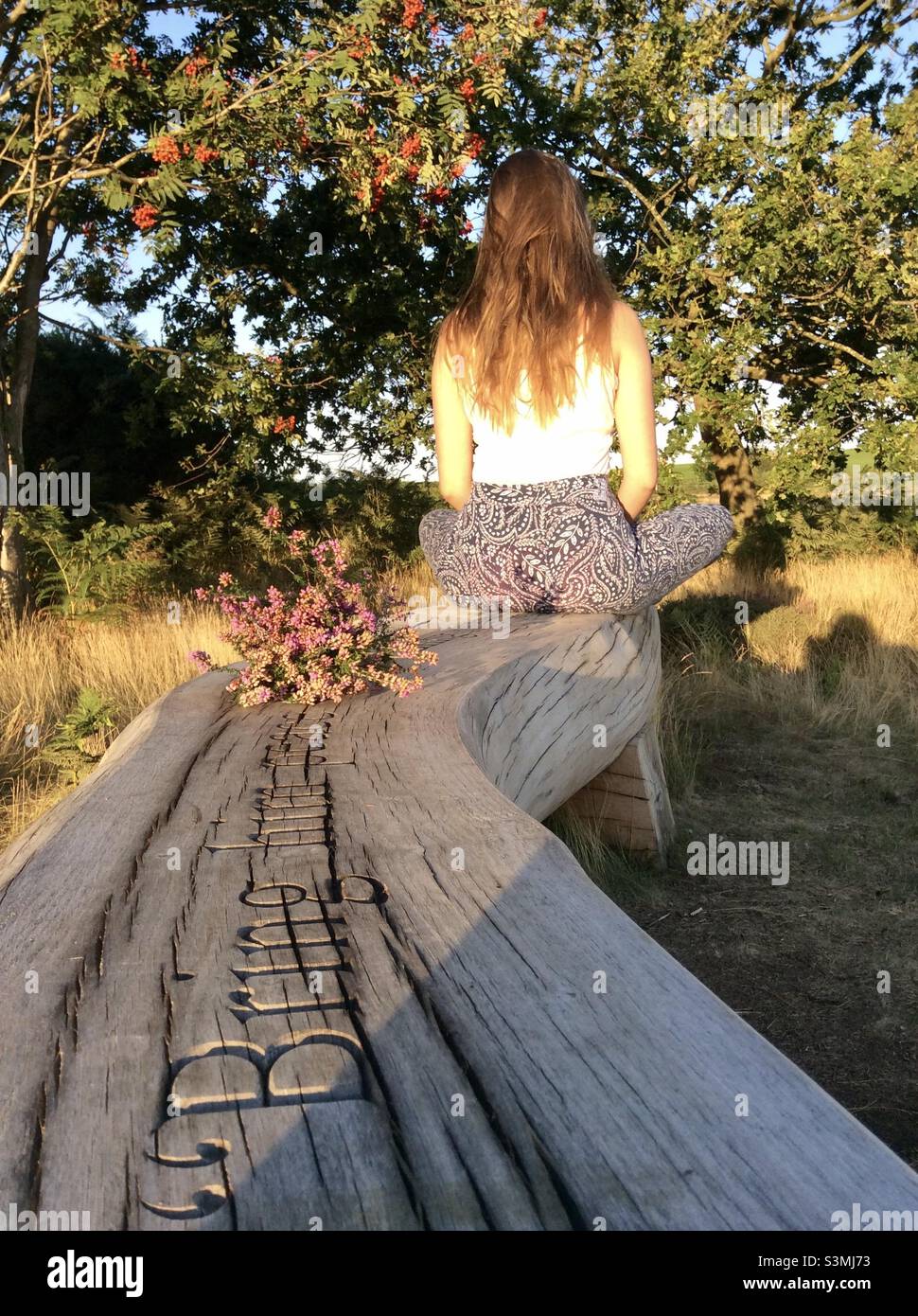 Girl sitting in the sunshine on a wooden bench, engraved with the words, “Bring him peace”, with a bunch of picked pink heather flowers Stock Photo