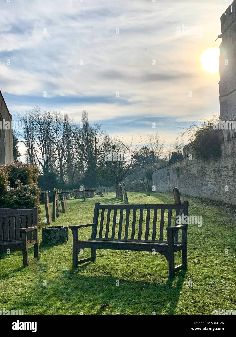 The low winter sun sits behind a stone building, setting shadows over the graveyard. A contemplation bench sits in the foreground. No people - Smartphone Captured Stock Image
