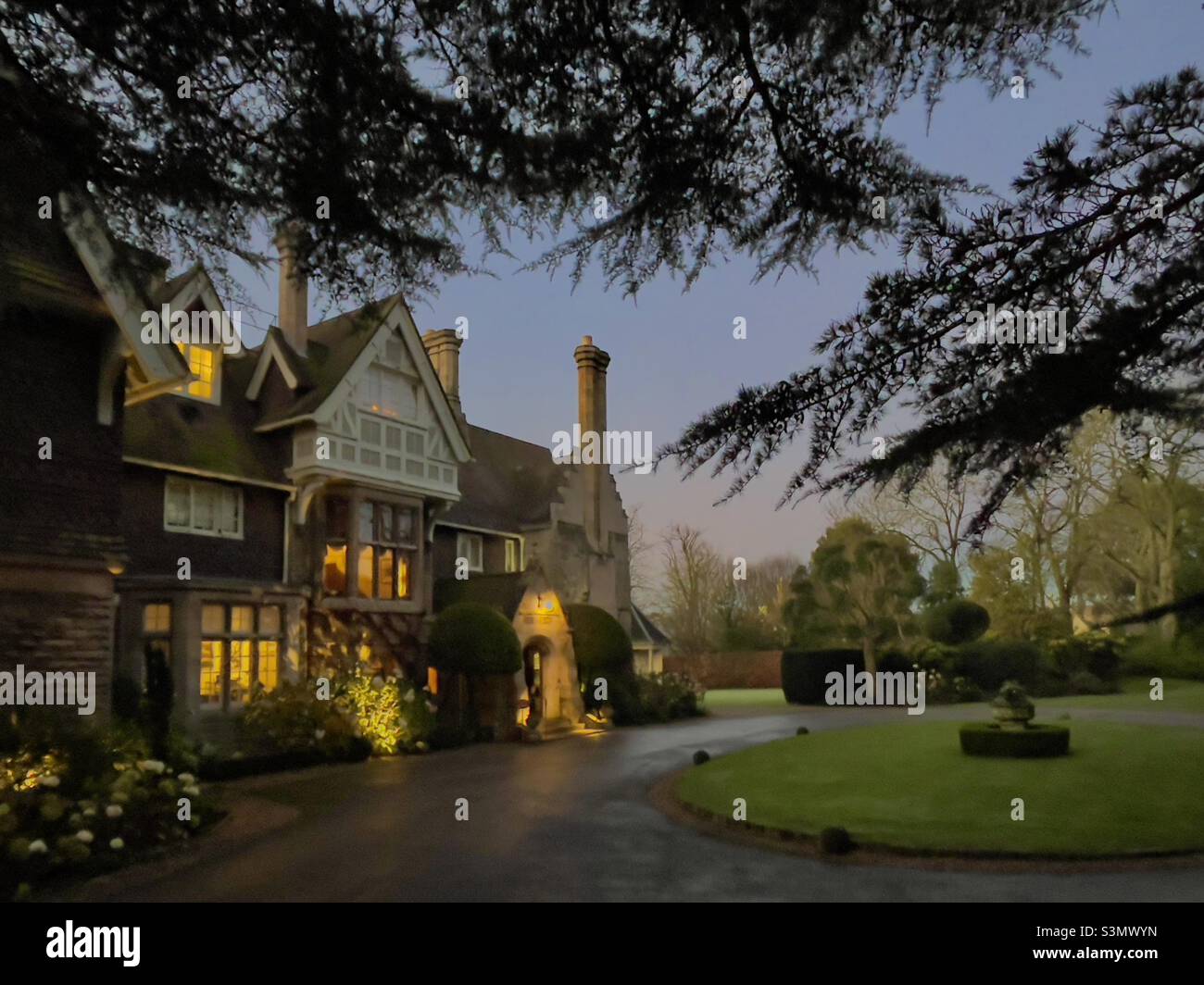 The exterior of a cosy country house hotel early on a winter morning, the lights still on and a cedar tree in the foreground - Smartphone Captured Stock Image