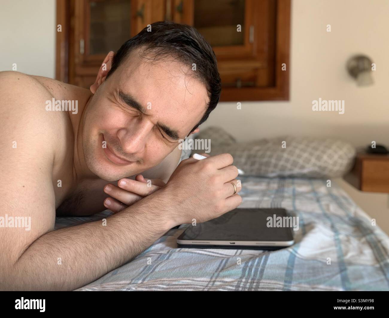 Photophobia. Young adult man working on tablet while laying still in bed closes his eyes because of the sunlight that entered when a door was opened. - Smartphone Captured Stock Image