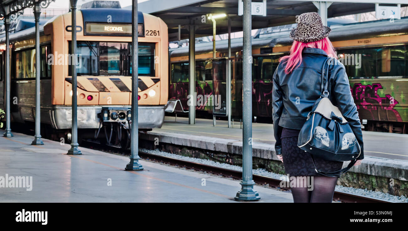 A woman dressed in black, with bright red hair and an animal print hat, faces an approaching train arriving at the station - Smartphone Captured Stock Image
