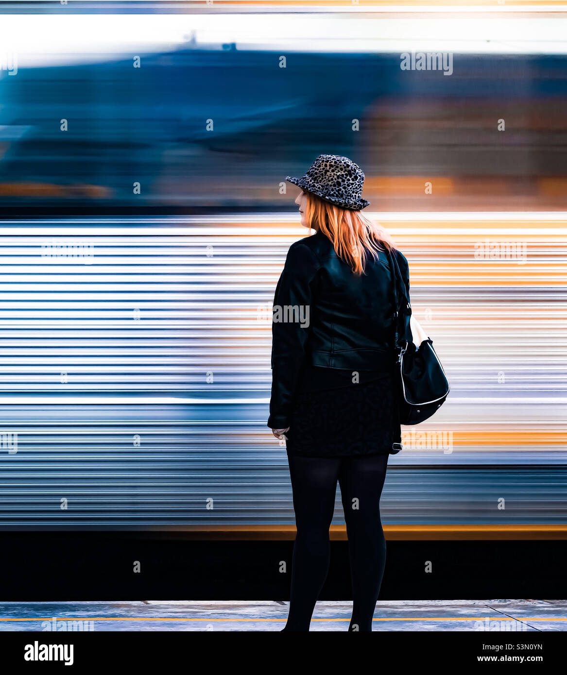 The metallic carriage of a train whizzes past a woman on the platform, her orangey yellow hair matches stripes on the train - Smartphone Captured Stock Image