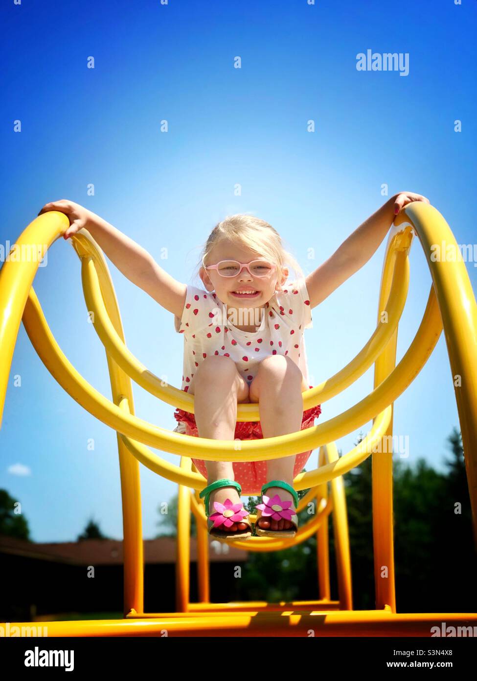 Little blonde girl with a big smile wearing glasses playing on playground equipment Stock Photo