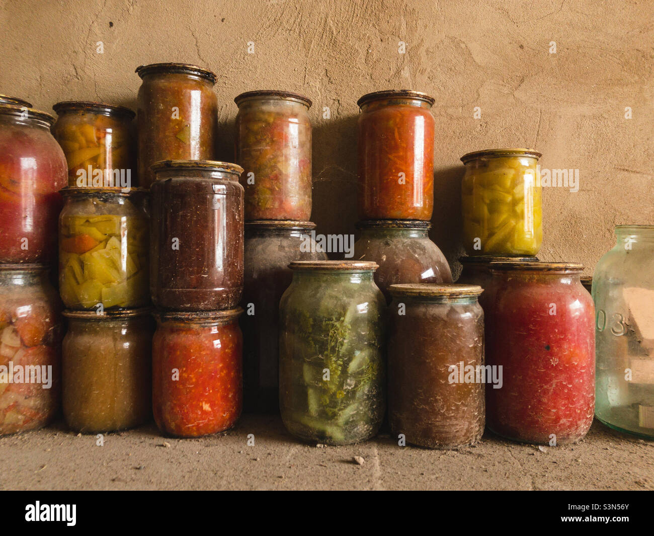 Canned vegetables, fruits and salads in glass jars under an iron lid stand on a dusty floor in a cellar in a garage. Stock Photo