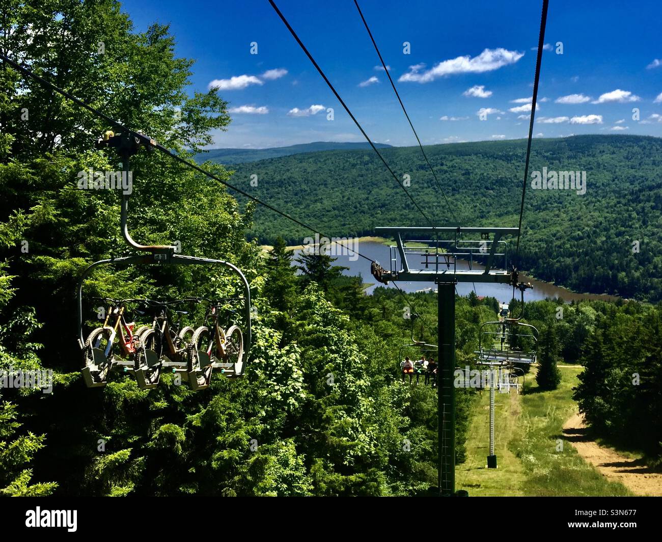 Riding down a ski lift through a forest of green trees toward a lake in summer with bicycles and unidentifiable people riding up Stock Photo