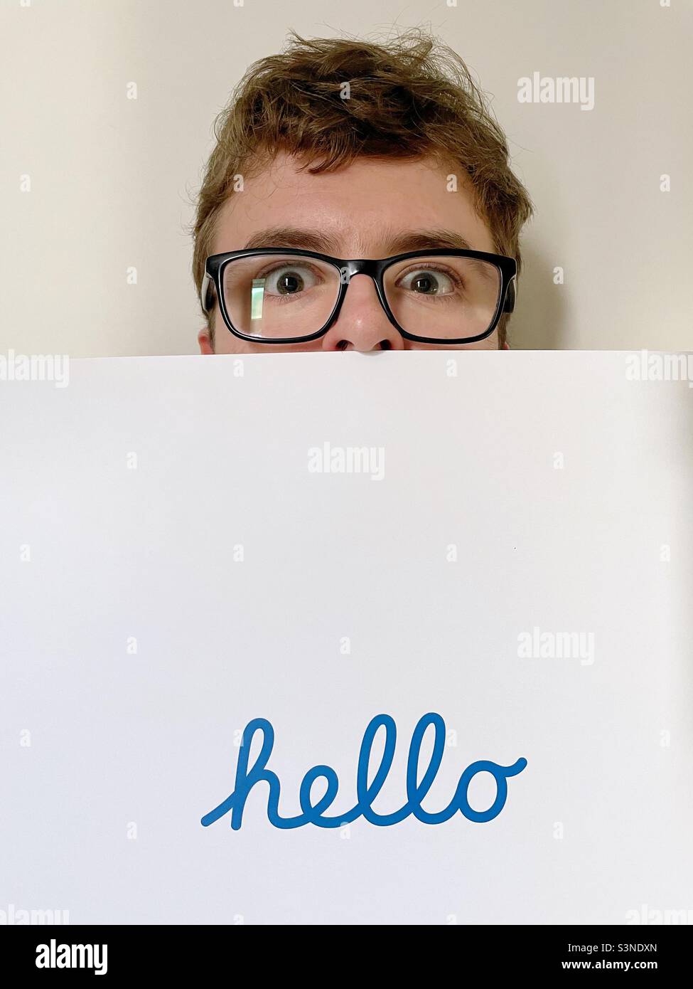 A shy and uncertain young male hiding behind a background with the word hello written in text during an introduction Stock Photo