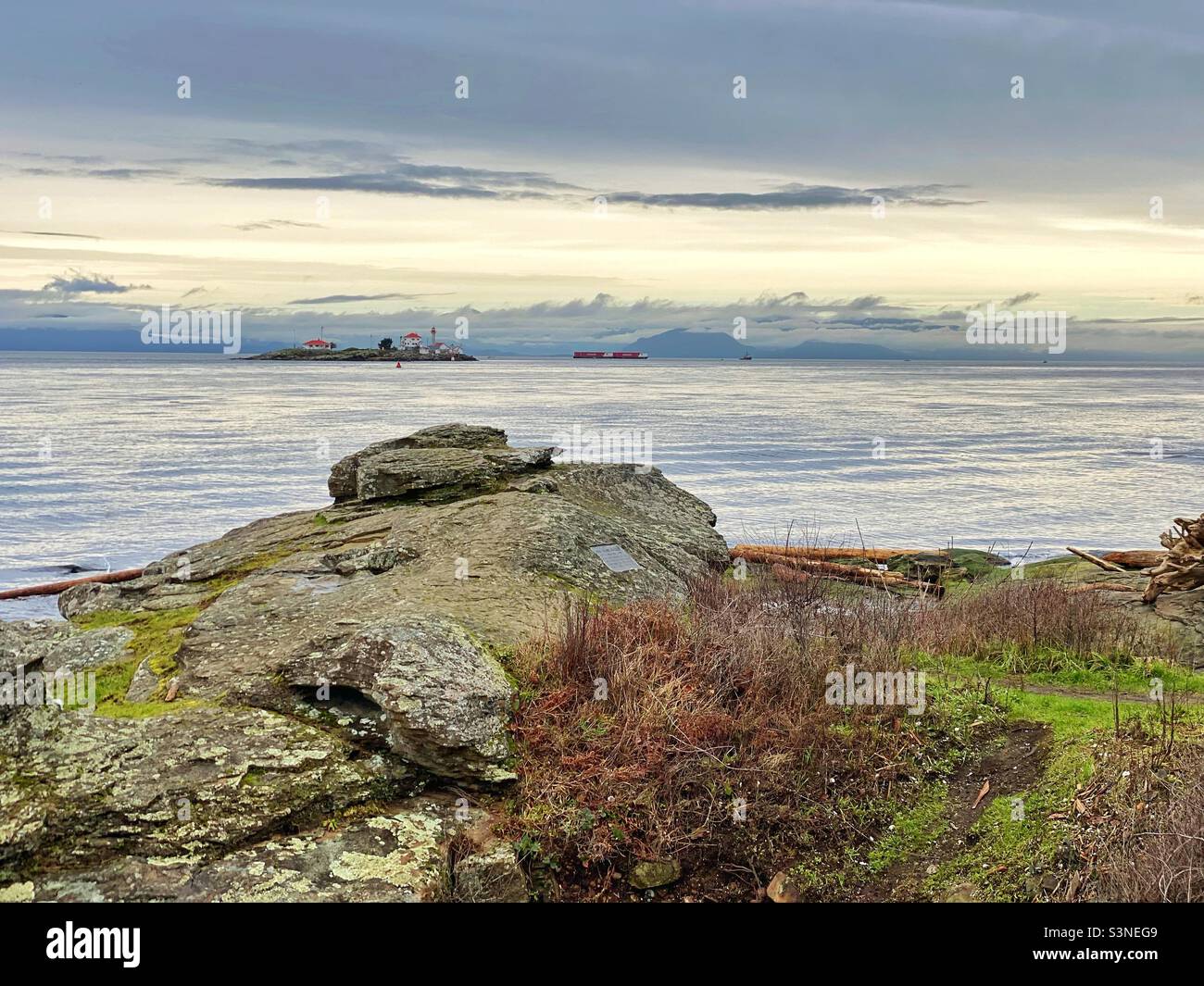 From a rocky point on a beach in British Columbia‘s Gulf Islands, the view is of a lighthouse in the Strait of Georgia and in the distance, tugboat towing two barges. Stock Photo