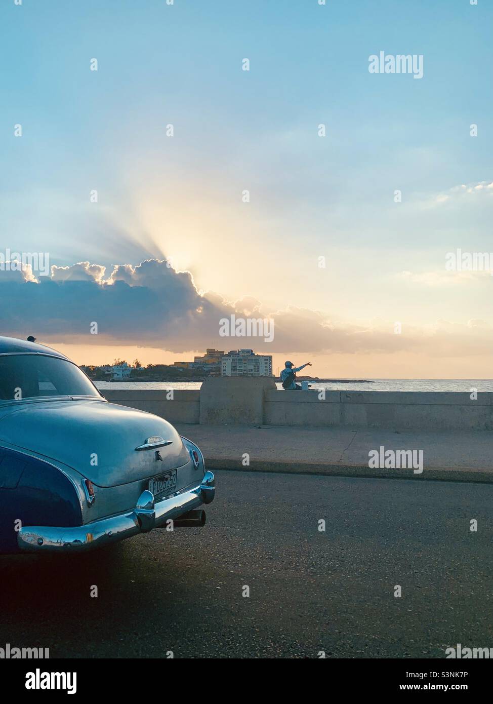 Havana February 3, 2022. Cubans Lifestyle. Man sitting in El Melecon, rear of Old Classic american car on the coastland avenue. Vedado, Havana, Cuba - Smartphone Captured Stock Image