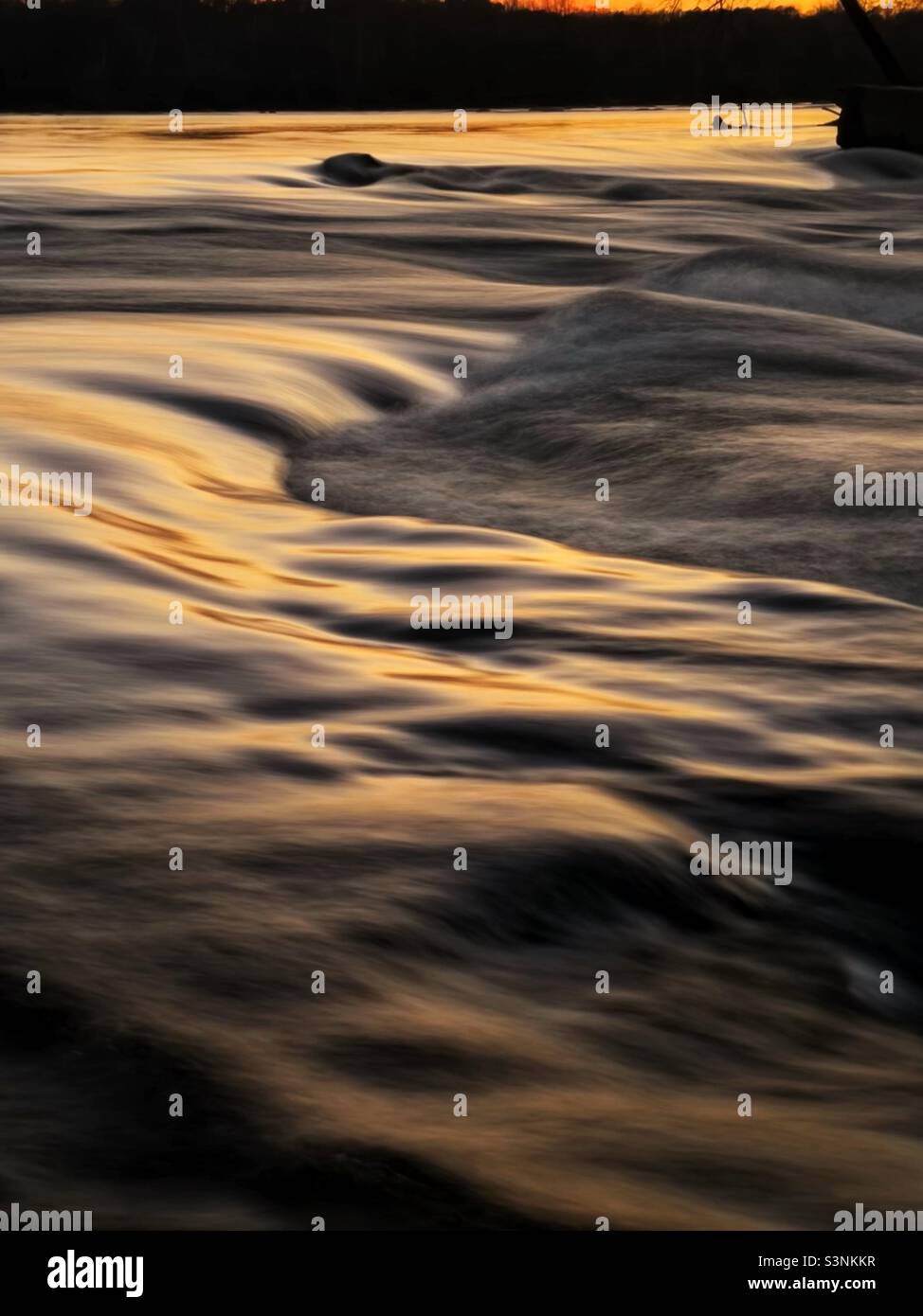 Smooth river waves flowing over rocks with an orange sunset reflected in the water. Stock Photo