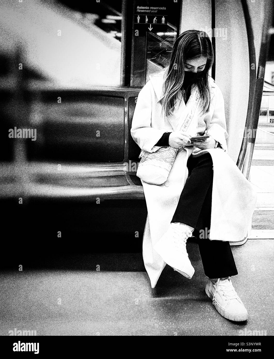 A young lady in an elegant long cream coat sitting on an underground train is checking her mobile phone. The photo is in black and white. Stock Photo
