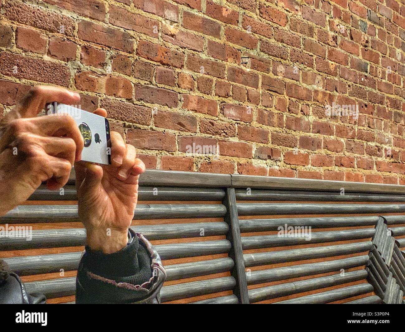 A man holding an iphone to take a picture against the bricks. - Smartphone Captured Stock Image
