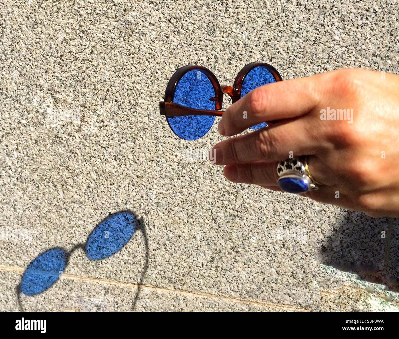 The shadow of a pair of blue sunglasses is cast onto a wall. The sunglasses are held in a lady’s hand. She is wearing an ornate silver ring in which a blue stone is set. - Smartphone Captured Stock Image