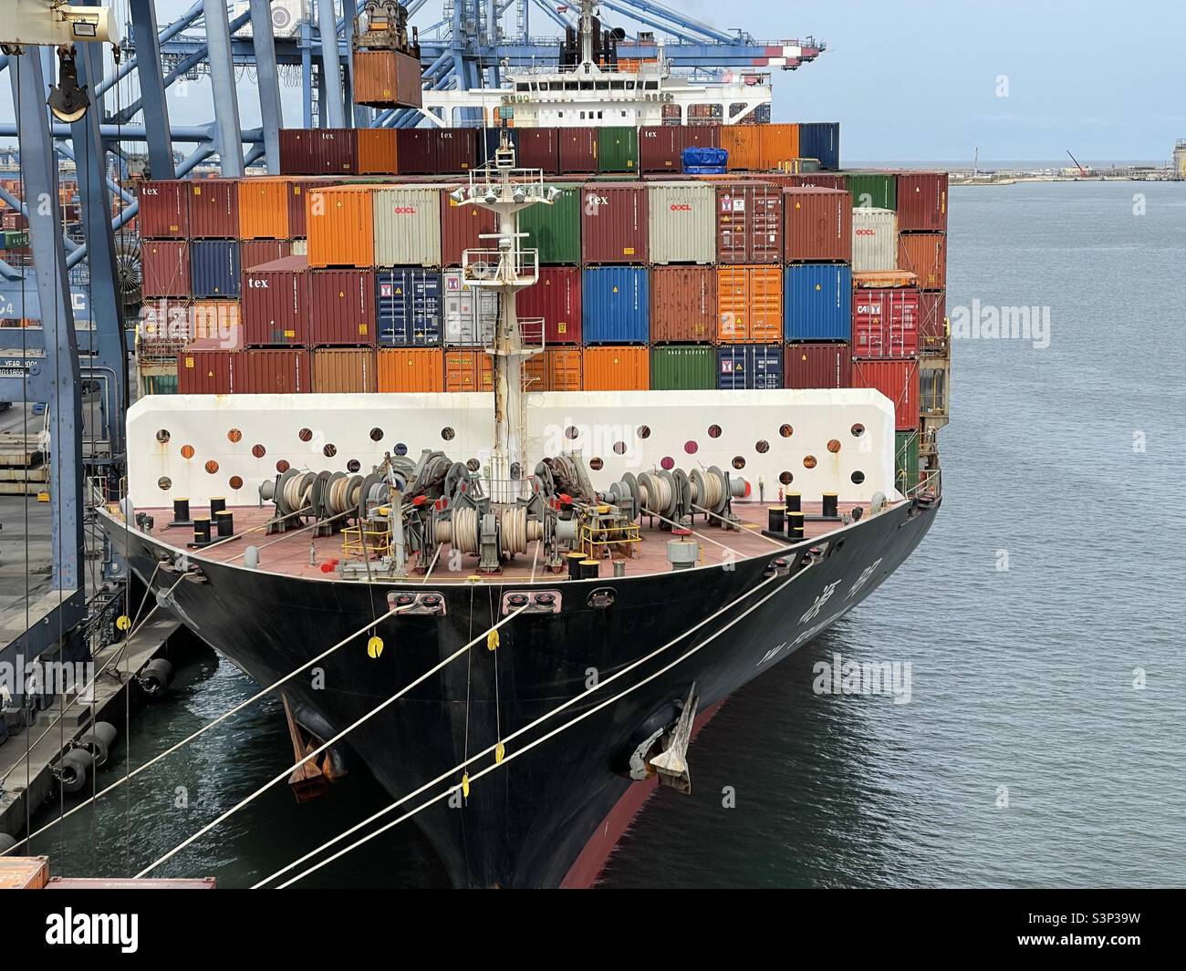 Forward mooring station of big loaded container  vessel in port terminal. Ship is fasted with ropes to the pier and is under cargo operation making by stevedores which using gantry cranes. Stock Photo